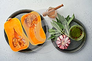 Halves of raw organic butternut squash with sage leaf, multicolored pepper garlic, honey, salt and pepper on a white slate, stone