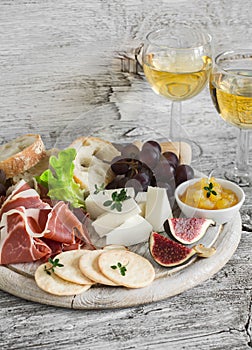 Ham, cheese, grapes, figs, nuts, bread ciabatta, cracker, jam on white wooden board and two glasses of white wine on bright wooden