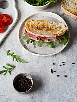 Ham, tomato and arugula sandwich on toasted bread slices on a white plate and other ingredients on a gray background