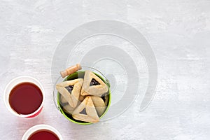 Hamantashen cookies or Aman ears, triangular cookies with poppy seeds in colored bucket on gray background and hot tea