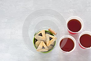 Hamantashen cookies or Aman ears, triangular cookies with poppy seeds in colored bucket on gray background and hot tea in paper cu