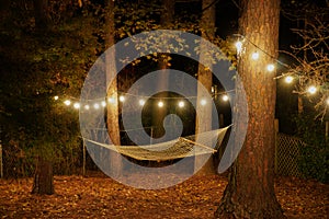 A hammock is suspended between two trees in a backyard with cafe lights strung up; romantic nighttime setting