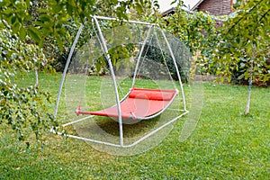 Hammock swing in metal frame with nobody on green lawn in backyard near house cottage. Rest relax relaxation alone on Red hammock