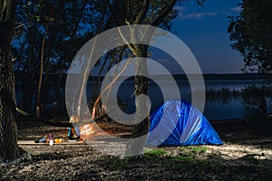 Hammok and girl is sitting near bonfire. Blue Camping Tent Illuminated Inside. Night Hours Campsite. Recreation and Outdoor. Lake