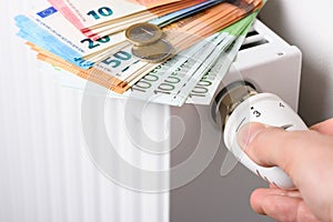 Hand adjusting the valve knob of heating room radiator temperature thermostat with stack of euro money banknotes and coins on it