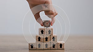 Hand arranges a wooden block with a dartboard icon stacked in a pyramid shape. Goal Achievement and Purposefulness, the challenge photo