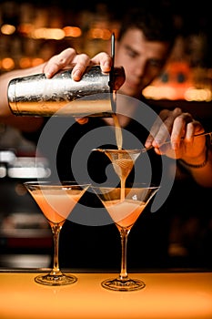 hand of bartender holds shaker with strainer and pours cocktail into martini glass.