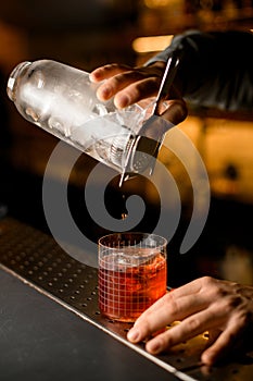 hand of bartender neatly holds mixing cup with strainer while pouring cold cocktail into glass
