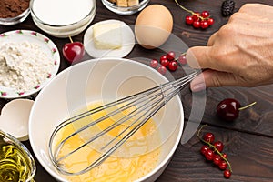 Hand beats eggs with a whisk in white bowl. Berries, flour, butter on table