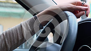 Hand of a beautiful white woman in beige blouse with watch and ring holds a black steering wheel. Hand of driver lies on top