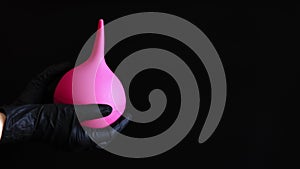 A hand in a black latex glove holds a large pink enema on a dark background