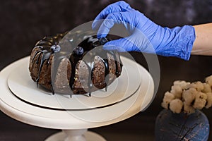 hand in a blue glove decorates with blueberries a chocolate cupcake with icing on a white plate next to a vase with a cotton