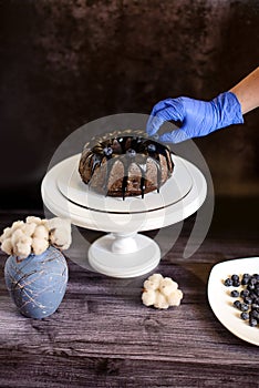 a hand in a blue glove decorates with blueberries a chocolate cupcake with icing on a white plate next to a vase with cotton