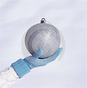 Hand with blue knit glove holding beautiful, big, sparkling, silver Christmas ornament with white snow background