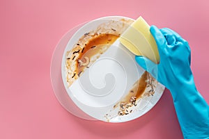 Hand in blue rubber glove hold yellow cleaning sponge to clean up and washing food stains and dirt on white dish after eating meal