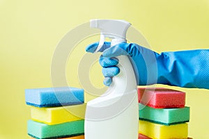 Hand in a blue rubber glove holds a spray bottle on a yellow background. Multi-colored cleaning sponges.