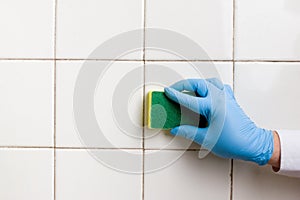 Hand in a blue rubber glove holds a yellow-green microfiber cleaning sponge, washing a white tile wall