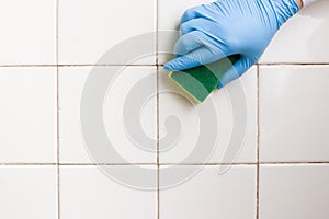 Hand in a blue rubber glove holds a yellow-green microfiber cleaning sponge, washing a white tile wall