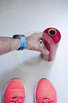 A hand with a blue sports smartwatch picks up a red water bottle from the floor during a break in a workout session at the gym,