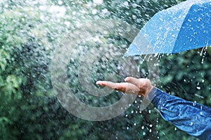 Hand and blue umbrella under heavy rain against nature background. Rainy weather concept
