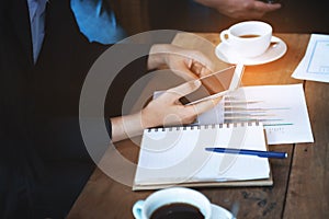 Hand of business asian woman use smart mobile phone contact client in group meeting in coffee cafe with note book and and paper of