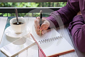 hand of business man holding pen writing on notebook paper. front of him have coffee cup with coffee and smartphone putting on