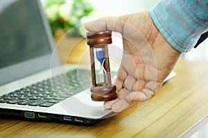 Hand of business man is holding wooden hourglass on laptop computer