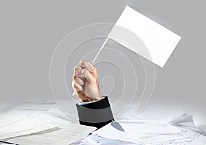 Hand of businessman emerging from loaded paperwork desk holding white flag