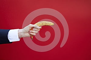 Hand of a businessman holding a banana as if shooting from a gun, with red background