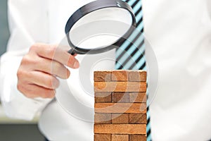 Hand of businessman holding glass magnifying over wooden blocks tower. Concept of risk analysis and business management.