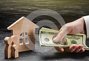 The hand of a businessman stretches a wad of money to the family figures near the wooden house. Providing a mortgage loan