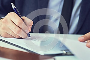 Hand of businessman in suit filling and signing with blue pen partnership agreement form clipped to pad closeup