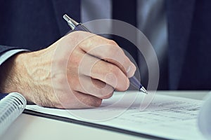 Hand of businessman in suit filling and signing with blue pen partnership agreement form clipped to pad closeup