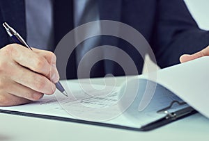 Hand of businessman in suit filling and signing with blue pen partnership agreement form clipped to pad closeup