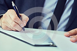 Hand of businessman in suit filling and signing with blue pen partnership agreement form clipped to pad closeup