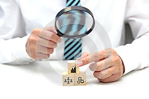 Hand of businessperson looking at wooden blocks with various icon symbols through magnifying glass.