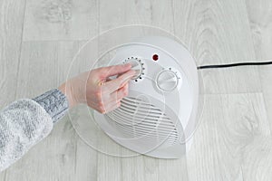 The hand of  caucasian woman turns on a fan heater on the floor of the room