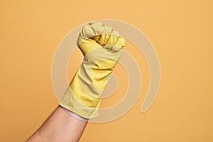 Hand of caucasian young man with cleaning glove over isolated yellow background doing protest and revolution gesture, fist