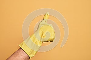 Hand of caucasian young man with cleaning glove over isolated yellow background doing successful approval gesture with thumbs up,