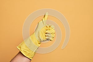 Hand of caucasian young man with cleaning glove over isolated yellow background doing successful approval gesture with thumbs up,