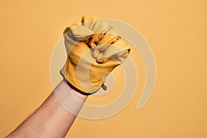 Hand of caucasian young man with gardener glove over isolated yellow background doing protest and revolution gesture, fist