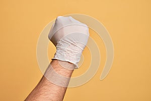 Hand of caucasian young man with medical glove over isolated yellow background doing protest and revolution gesture, fist