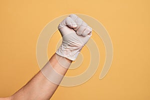 Hand of caucasian young man with medical glove over isolated yellow background doing protest and revolution gesture, fist