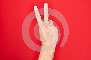 Hand of caucasian young man showing fingers over isolated red background counting number 2 showing two fingers, gesturing victory