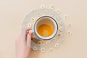 Hand with Ceramic Mug of chamomile tea with flowers on a beige background, top view with copy space for your design