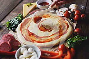 Hand of the chef smears tomato sauce on pizza dough, next to the ingredients for Margarita