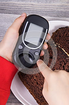 Hand of child holding glucose meter, sweet chocolate cake. Checking sugar level during diabetes