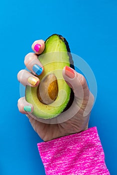 A hand with colourful nail polish holding a cut ripe avocado.