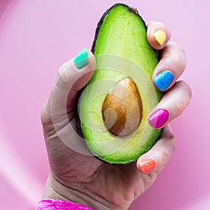 A hand with colourful nail polish holding a freshly cut avocado half.