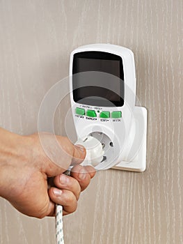 Hand connects plug with wattmeter socket on wall, for measuring electricity costs in device